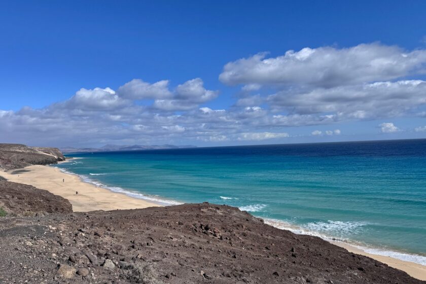 Sandstrand und türkisfarbenes Meer unter einem steinigen Vulkanhang auf Fuerteventura
